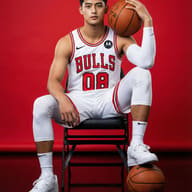 A young man in a white Chicago Bulls jersey sits on a chair against a red backdrop, holding one basketball and resting his foot on another. He wears white sneakers and athletic gear.