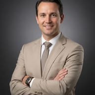 Professional portrait of a smiling man in a beige suit and tie, arms crossed against a dark grey background.
