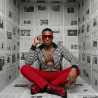 Gemini said Man in a striped jacket and red pants sits in a newspaper-lined room, wearing bold red sunglasses and boots. High-fashion, edgy editorial aesthetic.