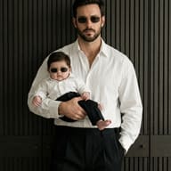 Man and Baby in Matching Sunglasses AI Photo Editing Prompt. AI generated fashion portrait of a bearded man holding a baby, both wearing white shirts and gold-rimmed sunglasses against a dark wood-paneled background.