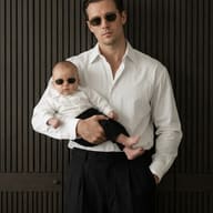 Man and Baby in Sunglasses AI Photo Editing Prompt. AI generated studio portrait of a stylish man holding a baby, both wearing white shirts and matching shades against a dark wood-slatted background.