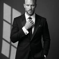 Man in Black Suit Black and White Photography Prompt. AI generated cinematic portrait of a sharp-dressed man in a black suit adjusting his tie against a shadowed studio background.