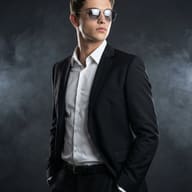 Man in Black Suit and Sunglasses AI Photo Editing Prompt. AI generated studio portrait of a stylish young man wearing a black suit and sunglasses posing against a dark smoky backdrop.