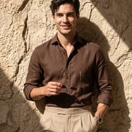 Man in Brown Linen Shirt Realistic Photography Prompt. AI generated cinematic portrait of a man wearing a brown linen shirt and beige trousers holding sunglasses against a textured stone wall.