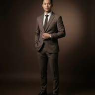 Professional full-body portrait of an East Asian man wearing a slim-fit brown suit, dark brown tie, and black dress shoes, standing against a solid brown studio backdrop.