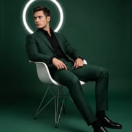 Man in Green Suit and Halo AI Photo Editing Prompt. AI generated studio portrait of a man in a dark green suit sitting on a white chair with a glowing ring background.