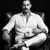Man in White Linen on Leather Chair AI Photo Editing Prompt. AI generated monochrome portrait of a man with a mustache in a white linen outfit sitting on a leather armchair and holding a fedora.