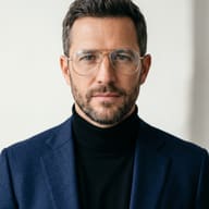 AI generated studio portrait of a bearded man wearing clear aviator glasses, a black turtleneck, and a navy blue blazer against a bright background.