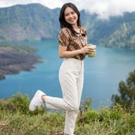Woman in Batik by Scenic Lake AI Photo Editing Prompt. AI generated cinematic lifestyle portrait of a smiling woman in a batik blouse and white trousers holding a matcha drink against a mountainous volcanic lake backdrop.